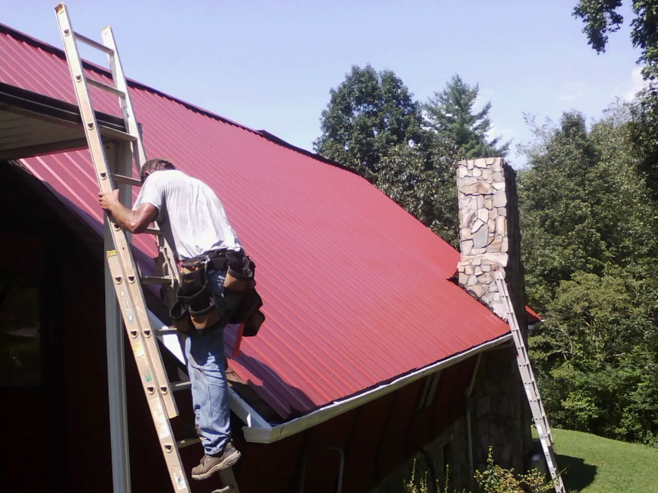 Roofer on a ladder beside a red metal roof and stone chimney — Soffit & Fascia Repair by Roofing Brothers in Markleysburg